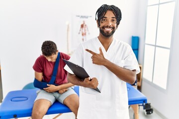 Wall Mural - Young hispanic man working at pain recovery clinic with a man with broken arm cheerful with a smile on face pointing with hand and finger up to the side with happy and natural expression