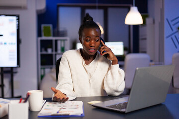 Stressed exhausted african american businesswoman talking with manager at phone discussing anuual company statistics late at night. Entrepreneur overworking at financial strategy in corporate office