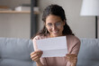 © fizkes - Smiling attractive young woman in glasses sit on sofa at home reading letter. Pleased female holds in hands sheet pleased by good news written in official document. Bank notice, happy client concept