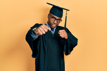 Young african american man wearing graduation cap and ceremony robe punching fist to fight, aggressive and angry attack, threat and violence