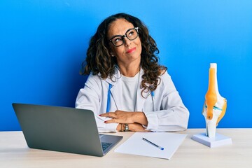 Poster - Beautiful middle age woman doctor at orthopedic clinic smiling looking to the side and staring away thinking.