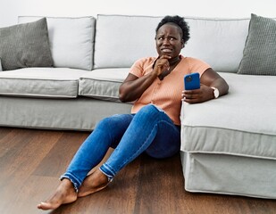 Sticker - Young african woman using smartphone sitting on the floor at home thinking worried about a question, concerned and nervous with hand on chin