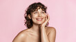 Smiling sensual woman with curly natural hair, glowing face and healhy skin, shiny body after shower. Happy girl touches her smooth and gentle face after using skin care product, pink background