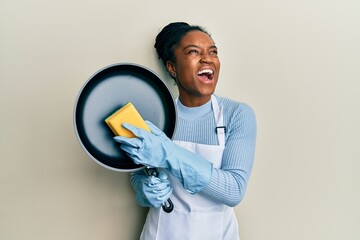 Canvas Print - African american woman with braided hair wearing apron holding scourer washing pan angry and mad screaming frustrated and furious, shouting with anger looking up.