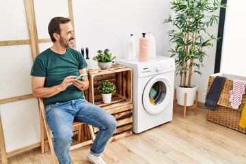 Wall Mural - Middle age hispanic man dong laundry using smartphone waiting at home.