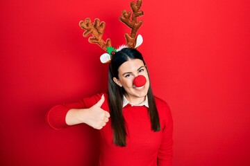 Canvas Print - Young hispanic woman wearing deer christmas hat and red nose doing happy thumbs up gesture with hand. approving expression looking at the camera showing success.