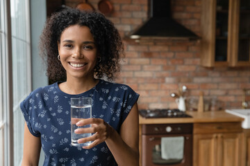 Wall Mural - Happy young African mixed race woman drinking fresh pure natural water in kitchen, holding transparent glass, looking, smiling at camera with toothy smile, keeping healthy habit. Head shot portrait