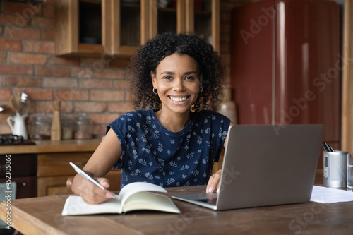 Portrait of happy African American student girl studying at laptop from home, making notes, looking at camera with toothy smile, attending virtual school class, online college lesson. Head shot