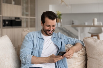 Wall Mural - Happy cellphone user getting good news, reading text message on smartphone, smiling at screen. Millennial man using mobile phone on couch at home, shopping online, chatting on social media