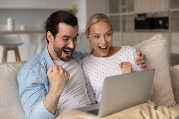 Wall Mural - Happy excited millennial couple with digital device celebrating success, achieve, win, getting good news. Shocked man and woman using laptop on couch, making winner yes gesture, shouting for joy