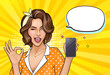 © redgreystock - Pop art girl holding mobile phone. Winking woman showing screen of new smartphone and OK sign. Pretty lady on yellow background with empty speech bubble. Vector illustration for digital advertisement.