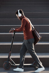 Wall Mural - Side view of young African man listening to music in headphones while pushing scooter against staircase