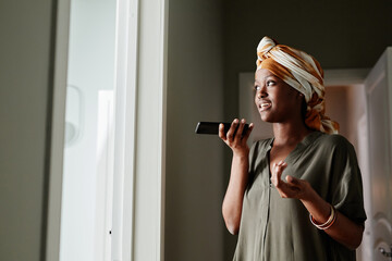 Wall Mural - Side view portrait of young African-American woman recording voice message while standing by window at home, copy space