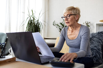 An older woman with short blonde hair sits on sofa in living room and works on laptop