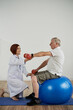 © DragonImages - Physical therapist asking senior man sitting on fitness ball to lift hand with dumbbell, rehabilitation concept