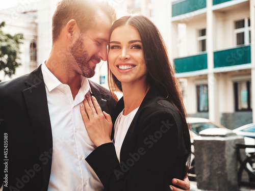 Beautiful fashion woman and her handsome boyfriend in suit. Sexy smiling brunette model in black evening dress. Fashionable couple posing in the street at sunset. Brutal man and his female outdoors