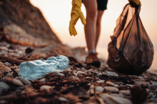 Close Up Of Blue Plastic Bottle Lying On The Beach. Close Up Of Volunteer's Hands With Plastic Bag Picks Up A Bottle. The Concept Of Earth Day And Envinmental Pollution