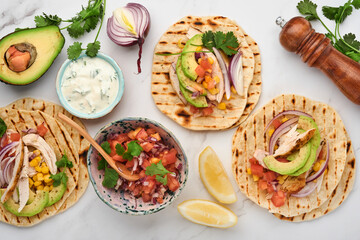 Mexican tacos with grilled chicken, avocado, corn kernels, tomato, onion, cilantro and salsa at white stone table. Traditional Mexican and Latin american street food. Top view.