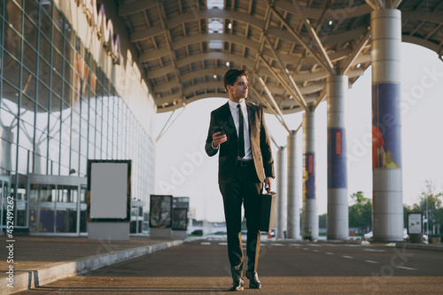 Bottom view full body traveler businessman man 20s in black suit go outside at international airport terminal with attache case use mobile cell phone book taxi order hotel Air flight business concept.