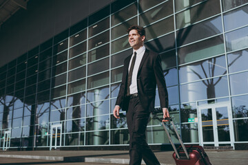 Wall Mural - Bottom view satisfied fun young traveler businessman man wear black dinner suit walk go outside at international airport terminal with suitcase valise crossing road Air flight business trip concept