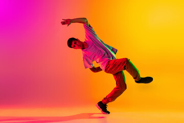 Wall Mural - Portrait of young stylish man, break dancing dancer training in casual clothes isolated over gradient pink yellow background at dance hall in neon light.