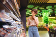 © ViDi Studio - Young minded woman in casual clothes shopping at supermaket greengrocery store buy choose meat sausages read ingredient shelf life prop up chin grocery cart inside hypermarke. Purchasing food concept
