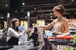 © ViDi Studio - Young smiling woman wear casual clothes mask shopping at supermaket stand at store checkout giving credit card to cashier pay inside hypermarket. People lifestyle purchasing gastronomy food concept.