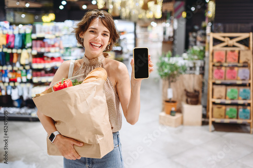 Young fun woman in casual clothes shopping at supermaket store with craft paper package with groceries show mobile cell phone blank screen workspace area inside hypermarket. Gastronomy food concept