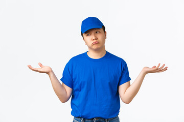 Indecisive clueless asian young delivery man in blue uniform, shrugging and looking away puzzled, spread hands sideways unaware, dont know. Courier standing perplexed over white background