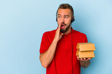 Wall Mural - Young caucasian delivery man with tattoos holding burgers isolated on blue background  is saying a secret hot braking news and looking aside