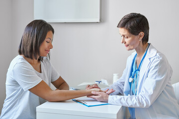 Wall Mural - Middle-aged woman doctor in white coat measuring pulse on patient wrist, checking heartbeat of young smiling afro american female visiting private clinic, professional physician doing health check up