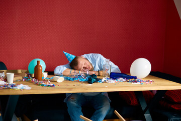 Man sleeping at table with blue cap in messy room after birthday party