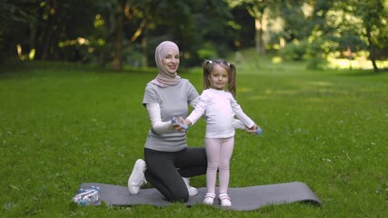 Wall Mural - Healthy lifestyle. Muslim mom in hijab helps her little daughter doing dumbbell exercises outdoors in the park. Happy kid raising dumbbells under supervision of her sporty mother