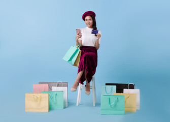 Beautiful Asian woman shopper sitting and carrying shopping bags with using credit card and mobile phone in hands on blue background.