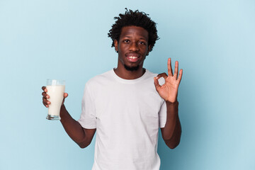 Young african american man holding a glass of milk isolated on blue background cheerful and confident showing ok gesture.