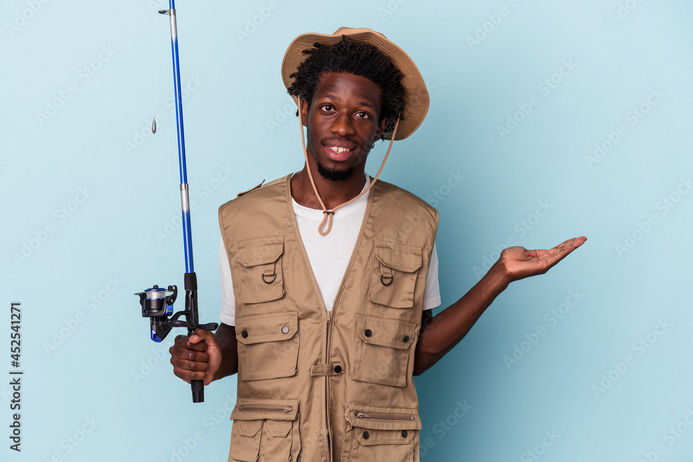 Young african american fisherman holding a rod isolated on blue background showing a copy space on a palm and holding another hand on waist. - obrazy, fototapety, plakaty 