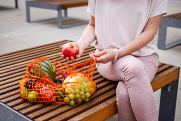 Wall Mural - Woman with reusable mesh bag sitting on bench in city. Resting at bench after shopping fruits in supermarket. Zero waste and plastic free concept