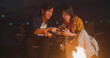 © tirachard - Young asia couple backpackers enjoying in camping at night near campfire on beach. Male and female traveler using smart phone at campsite. Outdoor activity, adventure travel, or holiday vacation.