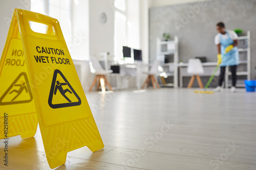 Close up plastic caution sign with figure that slips and falls warning us of wet slippery office floor. Black African American woman caretaker housekeeper cleaning modern room interior in background