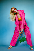 Fashionable Woman Wearing Trendy Fuchsia Color Suit. Orange Sunglasses, Holding Stylish Green Quilted Faux Leather Bag With Chunky Chain, Posing On Blue Background. Full-length Studio Fashion Portrait