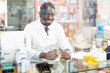 © JackF - Smiling african american male pharmacist working in a pharmacy makes important notes while sitting behind the counter