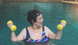 happy and healthy senior Asian woman  working out in the swimming pool with yellow dumbbells, smiling and looking away. Elderly sports , lifestyle.