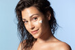 Pretty young smiling girl with sunscreen cream on perfect hydrated cheek skin, naked shoulders on blue studio background. Female advertising cosmetics with sun protecting, natural ingredients.