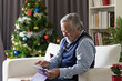 Senior Asian man with grey hair, beard, moustache using digital tablet on Christmas day while sitting on sofa at home. Merry Christmas and Happy Holidays. Christmas holiday celebration