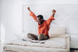 © wifesun - Joyful African-American guy in casual clothes and glasses puts hands up sitting near open laptop on comfortable bed at home