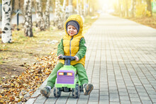Happy Little Boy Kid In Bright Yellow Vest Pushes Ride-on Toy While Walking On Paved Road In Sunny Autumn Park