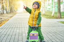 Sweet Little Boy In Bright Yellow Vest Points With Finger Standing Near Ride-on Toy Walking On Paved Road In Sunny Autumn Park