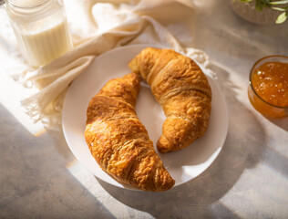 Breakfast table - two croissants, milk, jam. White silk tablecloth. Daylight illumination. Pastel shades. There are no people in the photo. Low angle view. Restaurant, hotel, recipe book.