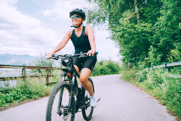 Portrait of a happy smiling woman dressed in cycling clothes, helmet and sunglasses riding a bicycle on the asphalt out-of-town bicycle path. Active sporty people concept image.