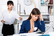 Frustrated woman sitting at office desk with disgruntled asian female boss behind